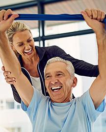An older man lifting weights while a woman supports him from behind
